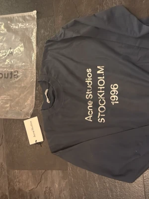 Mörkblå Acne Studios t-shirt - Snygg mörkblå t-shirt från Acne Studios med vit text 'Acne Studios STOCKHOLM 1996' på framsidan. Klassisk passform och rund halsringning. Perfekt för dig som gillar stilrena och trendiga plagg. Passar M/s
