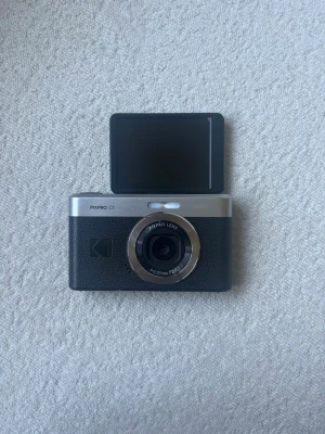 Kodak PIXPRO C1 digitalkamera - Snygg svart och silverfärgad Kodak PIXPRO C1 digitalkamera med en kompakt design. Kameran har en rund lins med silverring och en uppfällbar skärm för selfies eller vloggning. Perfekt för dig som vill ha en smidig kamera att ta med överallt Laddare medföljer såklart. Alla funktioner fungerar perfekt!