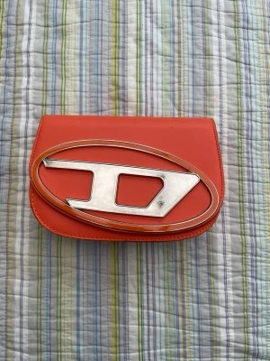 Orange kuvertväska med stor metallogo - Snygg orange kuvertväska i slätt konstläder med en stor, oval metallogo i silver på framsidan. Väskan har en stilren och modern design som sticker ut och passar perfekt till dig som vill ha något unikt.