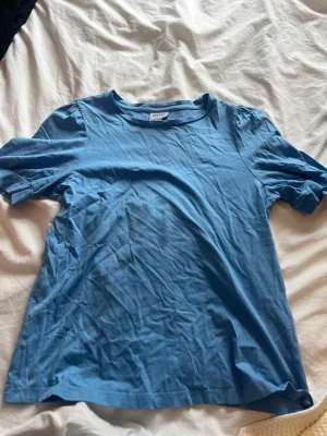 Blå basic t-shirt med puffärm - En enkel blå t-shirt med puffiga korta ärmar och rund halsringning. Tillverkad i mjuk bomull, perfekt för en avslappnad och stilren look. Passformen är normal och färgen är klarblå. Kommer inte till användning.