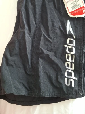 Svarta badbyxor från Speedo - Svarta badbyxor från Speedo med vit logga längs ena benet. Klassisk modell med ficka bak och praktisk dragsko i midjan. Perfekta för stranden eller poolen, tillverkade i snabbtorkande material.