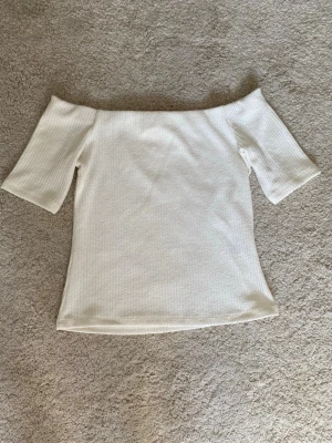 Vit offshoulder topp - Säljer en stilren vit offshoulder topp med ribbad struktur. Toppen har korta ärmar och en kroppsnära passform som ger en snygg siluett. 