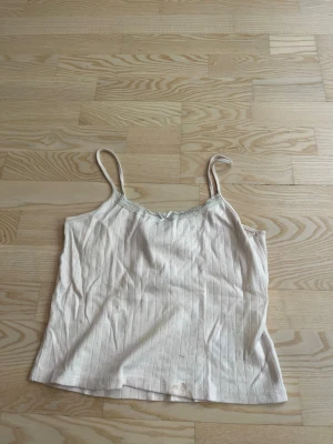 Beige ribbad linnetopp med spets - Söt rosa linnetopp med tunna axelband och ribbad struktur. Toppen har spetsdetalj vid urringningen och en liten rosett framtill. Perfekt för varma dagar eller som layering-piece.