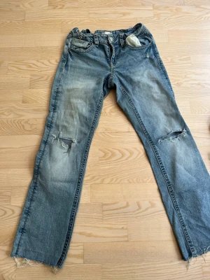 Blå slitna raka jeans - Ett par blå jeans med raka ben och slitningar på knäna samt fransiga benslut. Jeansen har klassisk femficksdesign och en något tvättad look. Perfekta för en avslappnad och trendig stil.