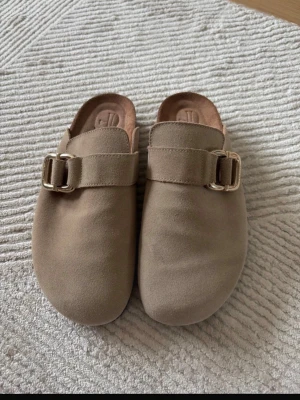 Beige slip-in tofflor med spänne - Stilrena beige slip-in tofflor i mockaimitation med bred rem och metallspänne. Tofflorna har rundad tå och öppen häl, samt en mjuk innersula för extra komfort. Perfekta för en avslappnad och trendig look hemma eller ute. Nypris 2000kr