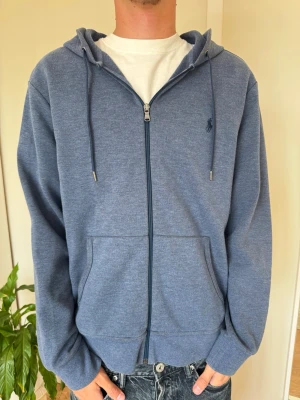Blå zip hoodie från Polo Ralph Lauren - Snygg blå hoodie med dragkedja från Polo Ralph Lauren. Klassisk modell med huva och dragsnören, två fickor fram och diskret logga broderad på bröstet. Tillverkad i mjukt sweatshirtmaterial som är skönt att ha på sig.