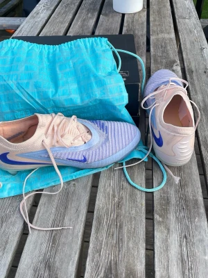 Nike phantom 6 ag elite low - Säljer ett par Nike fotbollsskor i en snygg ljusrosa och ljusblå färgkombination med blå Nike-logga på sidan. Skorna har snörning och en modern, strömlinjeformad design. Ovandelen är i textilmaterial som ger bra känsla på planen. Perfekta för dig som vill sticka ut!