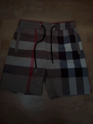 Burbbery shorts  - Snygga shorts med klassiskt rutigt mönster i beige, svart, vitt och rött. Shortsen har elastisk midja med svart snörning och sidofickor. Perfekta för sommaren och ger en trendig look.