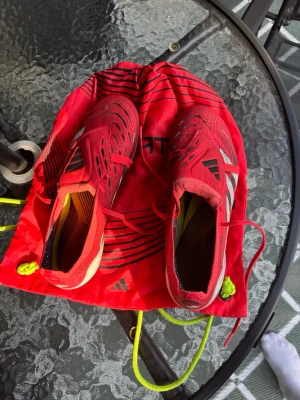Adidas Predator röda fotbollsskor - Säljer ett par röda Adidas Predator fotbollsskor med vita och svarta detaljer. Skorna har snörning, låg profil och är tillverkade i syntetmaterial för bra grepp och kontroll på planen. Medföljer matchande röd påse med Adidas-logga.