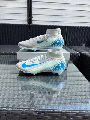 Nike mercurial superfly 10 - Snygga fotbollsskor i vitt med blå detaljer och högt skaft. Skorna har en stickad ovandel och snörning för bra passform. Skorna är i bra skick men det ända lilla problemet är att dem är lite smutsiga. Box och påse tillkommer! Ej original påse