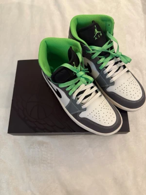 Air Jordan 1 Mid grå/vit/grön - Snygga Air Jordan 1 Mid sneakers i grått, vitt och svart med gröna detaljer och innersula. Skorna har både vita och gröna skosnören, perforerad tåbox och klassisk Jumpman-logga på plösen. Tillverkade i läder och textil för en clean och sportig look.