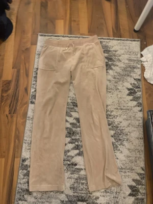 Beige mjukisbyxor med raka ben - Säljer ett par beige mjukisbyxor med raka ben och bred resår i midjan. Byxorna har snörning framtill, två stora fickor fram och två bakfickor med knappdetaljer. Materialet ser ut att vara mjukt och skönt, perfekt för chill dagar.