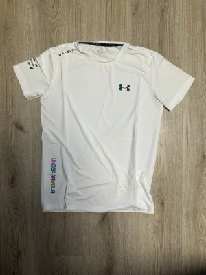Vit träningstopp från Under Armour - Säljer en vit kortärmad träningstopp från Under Armour med rund halsringning och logga på bröstet. Toppen har färgglad textdetalj på sidan och är tillverkad i ett lätt syntetmaterial som andas. Perfekt för dig som gillar sportig stil.