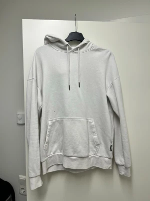 Vit hoodie med magficka och huva - Säljer en stilren vit hoodie med huva och dragsnören. Hoodien har en stor magficka framtill och ribbade muddar vid ärmslut och nederkant. Perfekt för en avslappnad och trendig look.