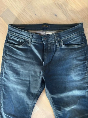 Mörkblå jeans från Jack & Jones - Snygga mörkblå jeans från Jack & Jones i modell regular Clark med slitningar och kontrastsömmar. Klassisk femficksmodell och läderpatch bak i midjan. Jeansen är tillverkade i ett mjukt denimtyg med stretch som ger en skön känsla. Mått: midja 41, innerben 72, ytterben 98. Storlek 31/30. 