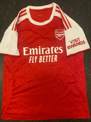 Arsenal matchtröja Saliba #2 röd/vit - Snygg Arsenal matchtröja i rött och vitt med Saliba #2 på ryggen. Tröjan har korta ärmar, klassiskt mönster och tryck med Emirates och Visit Rwanda. Perfekt för fotbollsfans som vill visa sitt stöd på läktaren eller hemma.