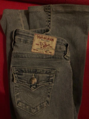 True Religion Joey Low Rise Flare Jeans stl 24 - Snygga ljusblå jeans från True Religion, modell Joey Low Rise Flare. Jeansen har låg midja, utsvängda ben och klassiska detaljer som kontrastsömmar och dekorativ bakficka med knapp. Perfekta för en avslappnad och trendig look. Nypris 1299kr storlek 24