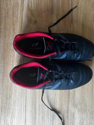 Svarta fotbollsskor Pro Touch - Snygga svarta fotbollsskor från Pro Touch med röda detaljer och svarta dobbar. Skorna har snörning och en låg profil, samt gråa detaljer på sidan. Insidan är vadderad med röd textil för extra komfort. Perfekta för konstgräs eller gräsplan.