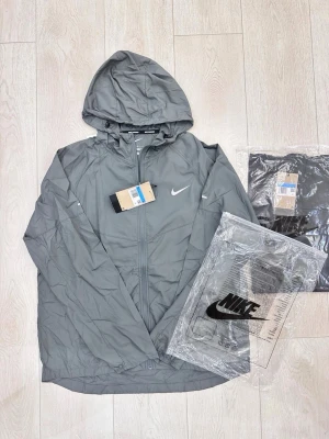 Grå vindjacka med huva från Nike Miller  - Snygg grå vindjacka från Nike med huva och dragkedja framtill. Jackan har en minimalistisk design med vit Nike-logga på bröstet och är tillverkad i lätt, vindtätt material. Perfekt för blåsiga dagar och enkel att matcha med olika outfits.