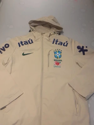 Beige Brasil CBF vindjacka med huva - Snygg beige vindjacka med huva och dragkedja framtill. Jackan har Brasilien CBF-logga, Nike-logga och flera sponsortryck i blått och grönt. Praktisk bröstficka med dragkedja och två sidofickor. Perfekt för dig som gillar sportig stil och fotboll.