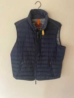 Mörkblå dunväst med dragkedja - Snygg mörkblå dunväst med hög krage och två fickor med dragkedja framtill. Västen har quiltad design och en gul detalj vid halsen. Perfekt för lager-på-lager och håller dig varm under kyliga dagar. Lagning under ena fickan se bildnr 6‼️