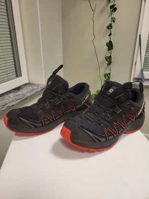 Salomon XA Pro Trailskor i Svart/Röd, Storlek 39 - Säljer ett par svarta kängor från Salomon med röda detaljer och snabbsnörning. Skorna har robust sula i rött och svart, samt slitstark ovandel i textil och syntet. Perfekta för dig som gillar outdoor och streetstyle.