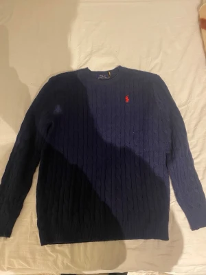 Mörkblå kabelstickad tröja Polo Ralph Lauren - Kabelstickad mörkblå tröja från Polo Ralph Lauren med rund halsringning och den klassiska röda logotypen broderad på bröstet. Tröjan har långa ärmar och ribbade muddar. Perfekt för dig som gillar stilrena och tidlösa plagg. Det står xl men det är M