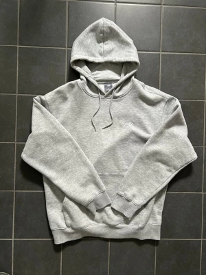 Grå hoodie från New Balance - Snygg grå hoodie från New Balance med klassisk huva och dragsnören. Hoodien har en liten diskret logga på bröstet, ribbade muddar vid ärmar och nederkant samt en stor magficka. Perfekt för en chill och sportig look.