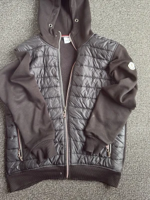 Svart cardigan med quiltad front - Snygg svart hoodie med quiltad front och släta ärmar. Dragkedja framtill, två fickor med dragkedja och ribbade muddar. Huva med metallöljetter och Moncler-logga på ärmen. Perfekt för en chill och stilren look.