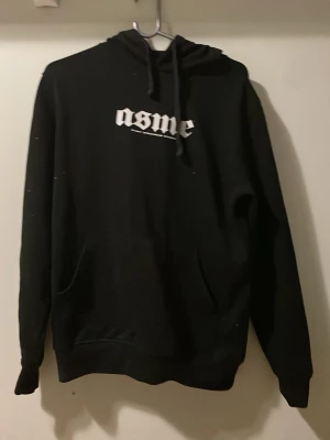 Exklusiv svart konsert hoodie med tryck - Säljer en svart hoodie med vit text 'asme' på bröstet och stort tryck på ryggen med texten 'The World Is Yours' och en jordglob. Hoodien har huva med dragsko och magficka. Perfekt för dig som gillar streetwear och vill sticka ut med coolt tryck.