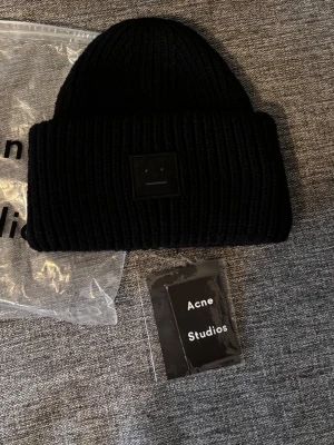 Svart ribbstickad mössa Acne Studios - Säljer en svart ribbstickad mössa från Acne Studios med den klassiska ansiktsloggan framtill. Mössan har bred uppvikt kant och är tillverkad i ett tjockt, mjukt material som håller dig varm. Perfekt accessoar för kalla dagar.