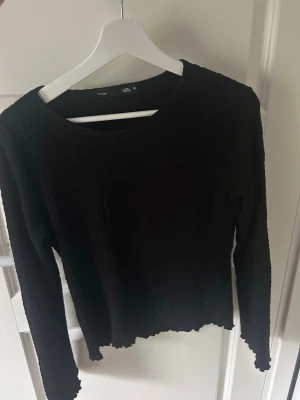 Svart långärmad topp med vågig kant - En svart långärmad topp med rund hals och vågiga kanter vid ärmslut och nederkant. Tunn och stretchig modell som sitter snyggt på kroppen och är enkel att styla till olika looks.