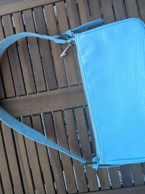 Ljusblå axelväska från Gina Tricot - Snygg ljusblå axelväska från Gina Tricot i croco-mönstrat konstläder. Väskan har en lång axelrem och stängs med dragkedja. Perfekt för att lyfta din outfit med en pop av färg och trendig struktur.