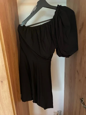 Svart oneshoulder klänning - Säljer en svart oneshoulder klänning med puffärm och draperad axel. Klänningen har en kort längd och är tillverkad i ett mjukt tyg som ger ett snyggt fall. Perfekt för dig som vill sticka ut med en unik siluett.