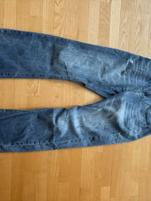 Jeans  - Säljer ett par blå jeans med slitningar och distressed detaljer. Jeansen har normal passform, klassisk femficksdesign och är tillverkade i bomull. Perfekta för en avslappnad och trendig look.