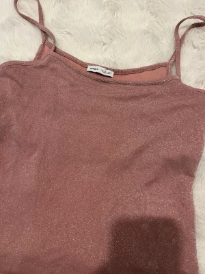 Glittrig rosa linnetopp med smala band - Säljer en glittrig rosa linnetopp med smala axelband. Toppen har en mjuk, skimrande yta och är perfekt för dig som vill sticka ut lite extra. Enkel och snygg design som passar till många olika stilar.