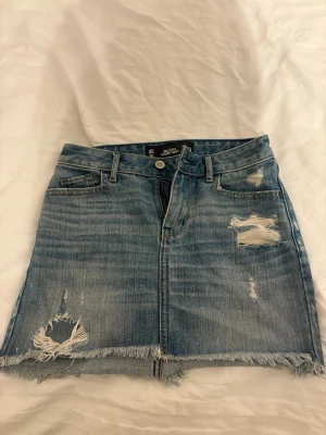 Hollister jeanskjol - Säljer en kort blå jeanskjol med slitna detaljer och fransig nederkant. Kjolen har klassiska fickor fram och bak samt bälteshällor. Perfekt för dig som gillar en avslappnad och trendig stil.