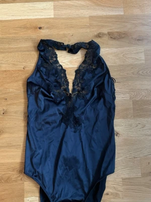 Svart spetsbody med djup urringning - Snygg svart body i satinliknande material med djup urringning och dekorativa spetsdetaljer framtill. Hög skärning vid benen och knäppning i nacken