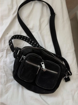 Noveller väska med nitar - Cool svart handväska i mockaimitation med två framfickor och silvriga dragkedjor. Väskan har en bred axelrem med nitar och justerbart spänne. Perfekt för dig som gillar en edgy och trendig stil.