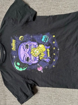 Svart Avengers Thanos t-shirt - Cool svart t-shirt från Pop! Tees med Marvel Avengers Infinity War-tryck. Stor färgglad bild av Thanos med Infinity Gauntlet och planeter i bakgrunden. 