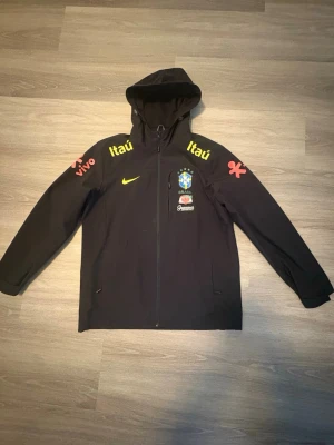 Svart Brasilien Nike träningsjacka - Svart träningsjacka med huva från Nike, prydd med Brasilien fotbollslandslagets emblem och flera sponsorloggor i gult och rött. Jackan har hel dragkedja framtill, två sidofickor och tryck på både bröst och ärmar. Perfekt för sportiga tillfällen.