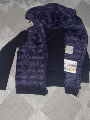 Marinblå Moncler cardigan - Marinblå pufferjacka från Moncler med quiltad front och huva. Jackan har stickade ärmar och muddar, dragkedja framtill samt två fickor med dragkedja. Klassisk Moncler-logga på ärmen och insida med serietidningsdetalj. Perfekt jacka nu inför höst. Nfc tag finns självklart. Om fler bilder önskas är det bara att höra av sig. 