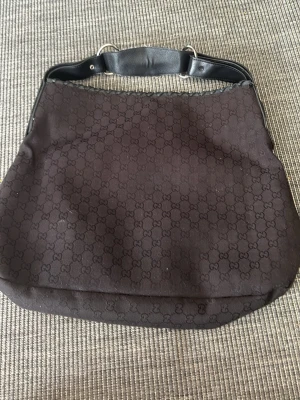 Svart axelremsväska från Gucci - Snygg svart handväska från Gucci med klassiskt GG-mönster över hela väskan. Väskan har en bred axelrem i svart läder med silverfärgade metalldetaljer. Perfekt för dig som gillar stilrena och tidlösa accessoarer.