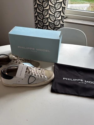 Philippe Model vita sneakers - Snygga vita sneakers från Philippe Model med beige mockadetaljer och svart häl. Skorna har klassisk låg profil, vita snören och diskret logga på sidan. Perfekta för en stilren och trendig look.
