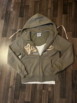 Grön zip-hoodie med Gällnö-tryck - Säljer en grön hoodie med dragkedja och stor huva. På bröstet står det 'Gällnö Stockholms Skärgård' i beige och vitt tryck. Hoodien har fickor framtill och ribbade muddar. Perfekt för chill dagar eller när du vill ha en avslappnad stil.