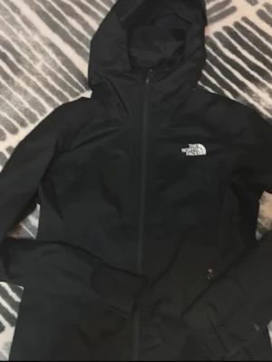 Svart vindjacka från The North Face - Snygg svart vindjacka från The North Face med huva och dragkedja framtill. Jackan har logga på bröstet och ryggen, samt smidig passform och tunnt material som skyddar mot vind. Perfekt för dig som vill ha en stilren och funktionell jacka.