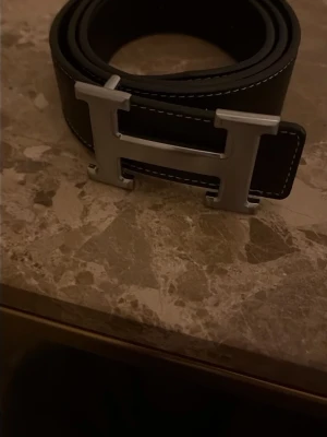 Svart bälte med stor H-spänne - Snyggt svart bälte med ett stort H-format metallspänne och vita sömmar längs kanterna. Bältet har en stilren och klassisk look som passar till många outfits. Perfekt accessoar för dig som vill ha något som sticker ut.