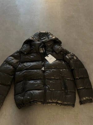  pufferjacka från Moncler - Snygg svart pufferjacka från Moncler med glansig finish och huva. Jackan har två dragkedjefickor framtill, diskret Moncler-logga på ärmen och dragkedja hela vägen upp. Perfekt för kalla dagar och riktigt trendig look. Storlek 5 altså xl men skulle säga att den sitter mer som L! 