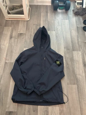 Stone Island Shell jacka - Snygg mörkblå vindjacka med huva och dragkedja framtill. Jackan har en bröstficka med dragkedja och ett svart tygmärke med gul och grön logga på vänster ärm. Resår vid ärmslut och justerbar nederkant med dragsko. Perfekt för blåsiga regniga och winter dagar. Med vänlig hälsning, Vincent OBS spännet är sönder