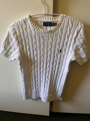 Kortärmad kabelstickad tröja från Polo Ralph Lauren storlek S - Klassisk kortärmad, kabelstickad tröja från Polo Ralph Lauren i en fräsch vit/off-white färg. Mörkblå polospelare broderad på bröstet. Material: bomull. Mycket fint skick. 